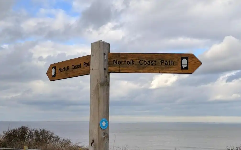 Wooden Norfolk Coast Path signpost overlooking the North Sea, with directional arrows displaying the National Trail acorn symbol beneath a cloudy coastal sky.