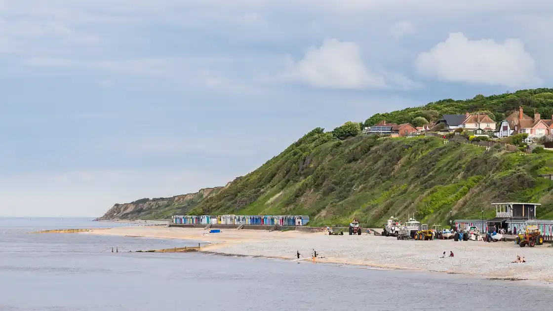 Overstrand Beach on the Norfolk coast, showing a sandy shoreline, colourful beach huts, fishing boats and steep green coastal cliffs with houses overlooking the North Sea.