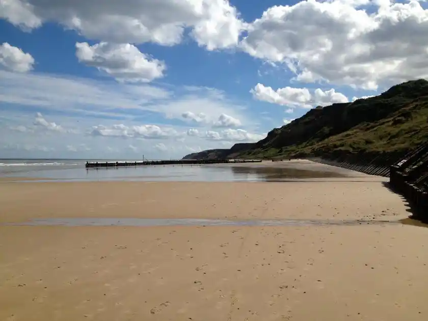 Wide sandy beach at Overstrand on the Norfolk coast at low tide, with timber groynes, steep coastal cliffs and a partly cloudy blue sky overlooking the North Sea.