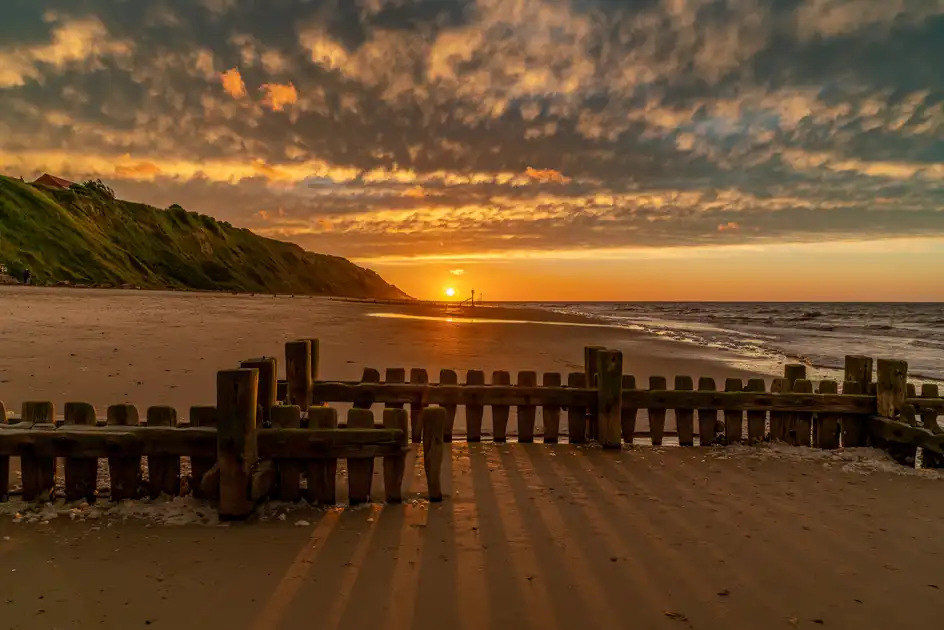 Golden sunset over Mundesley Beach on the Norfolk coast, with sandy shoreline, coastal cliffs, wooden sea defences and dramatic clouds above the North Sea.