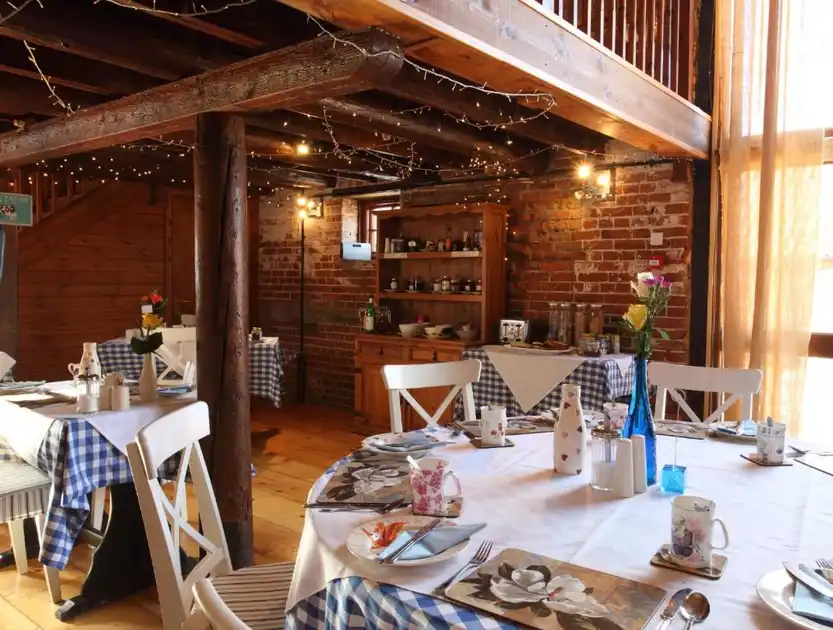 Breakfast dining room at Dairy Barns in Hickling, Norfolk, featuring exposed brick walls, timber beams, a mezzanine gallery, white wooden chairs and tables laid with blue checked tablecloths.
