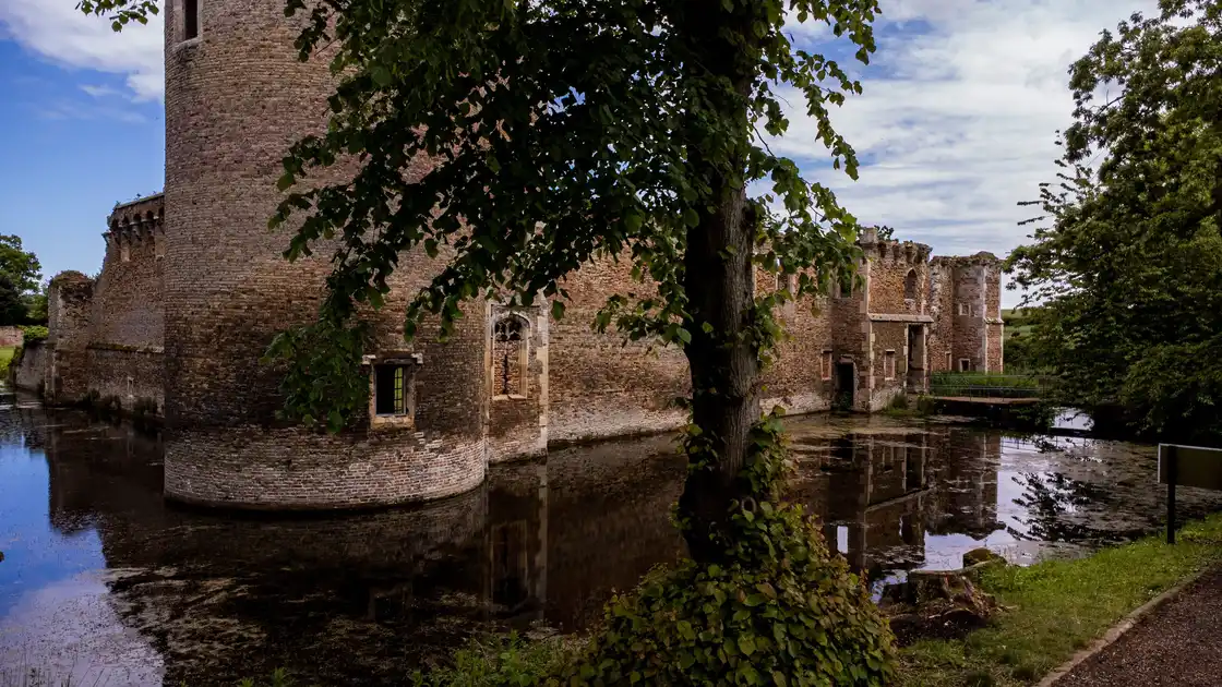 Medieval ruins of Caister Castle near Caister-on-Sea, Norfolk, showing the historic brick tower and fortified walls surrounded by a water-filled moat, with mature trees and reflections in the water.