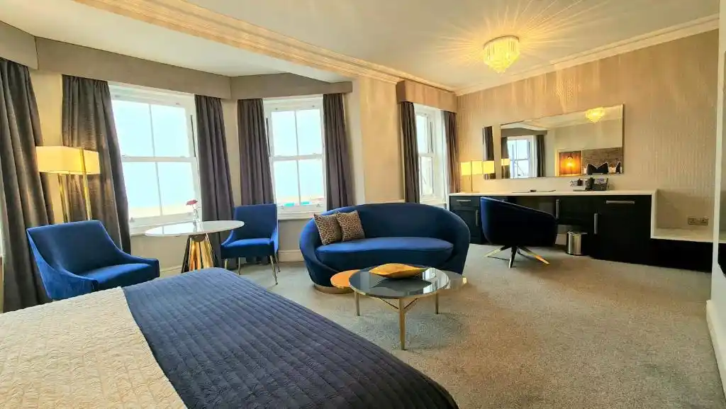 Spacious guest suite at Hotel Ocean in Great Yarmouth, Norfolk, featuring blue velvet sofas and armchairs, a large double bed, coffee table, dressing area and tall windows overlooking the seafront.