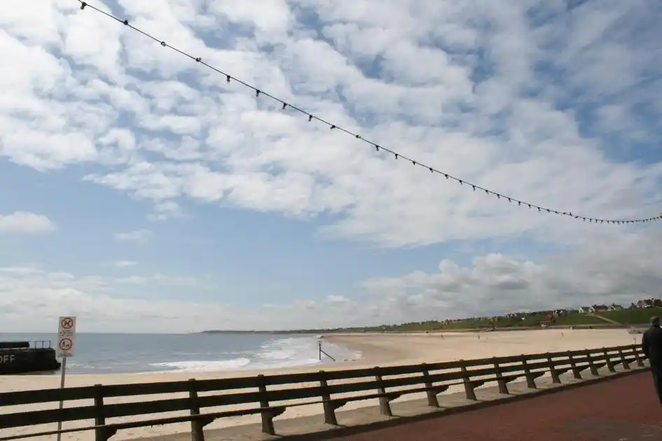 View along Gorleston Beach in Norfolk, showing the wide sandy shoreline, gentle waves, wooden railings beside the seafront promenade and coastal cliffs beneath a partly cloudy sky.