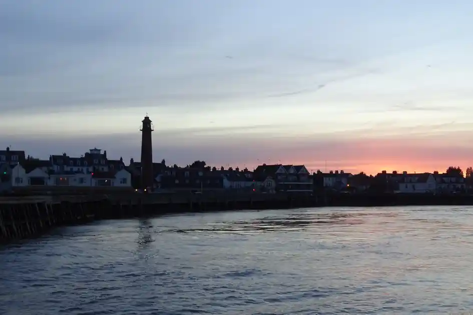View across the River Yare at Gorleston-on-Sea, Norfolk, showing the harbour lighthouse silhouetted against a pink sunset sky, waterfront buildings and reflections on the water.