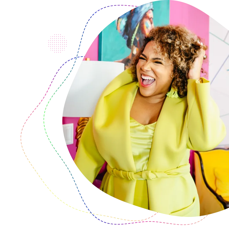 Smiling woman with curly hair wearing a bright yellow outfit, standing indoors with colorful artwork and furniture in the background.