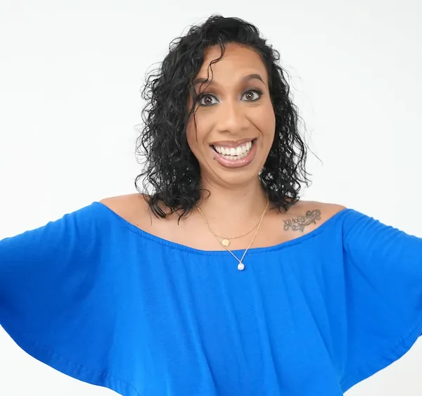 Woman with curly dark hair wearing a bright blue off-shoulder top, smiling broadly with arms open.