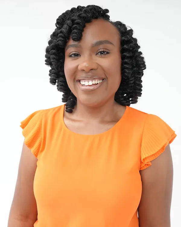 Smiling woman with curly black hair wearing an orange top with pleated short sleeves against a white background.