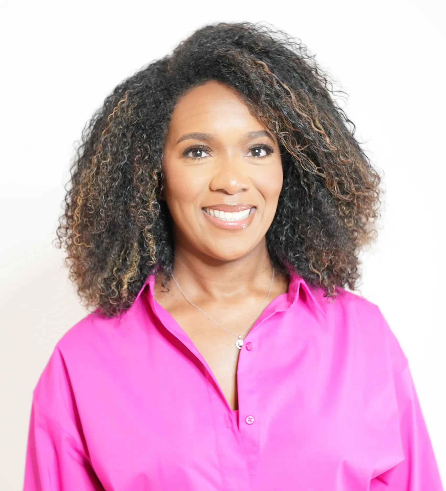 Smiling woman with curly hair wearing a bright pink shirt and a silver necklace against a white background.