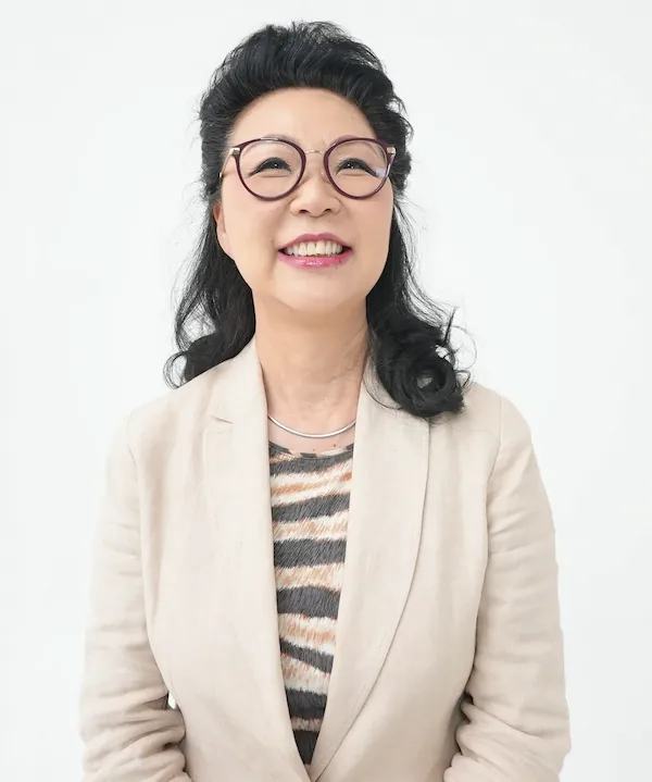 Smiling middle-aged Asian woman with black hair, wearing round glasses, a beige blazer, and a patterned top.