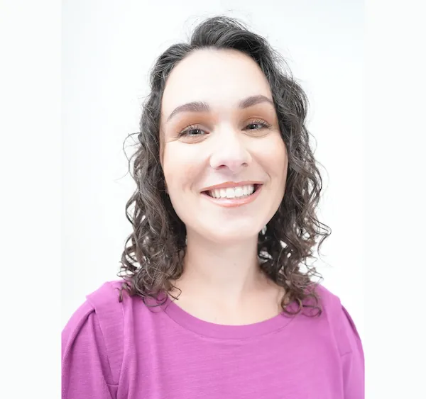 Smiling woman with curly dark hair wearing a purple top against a white background.