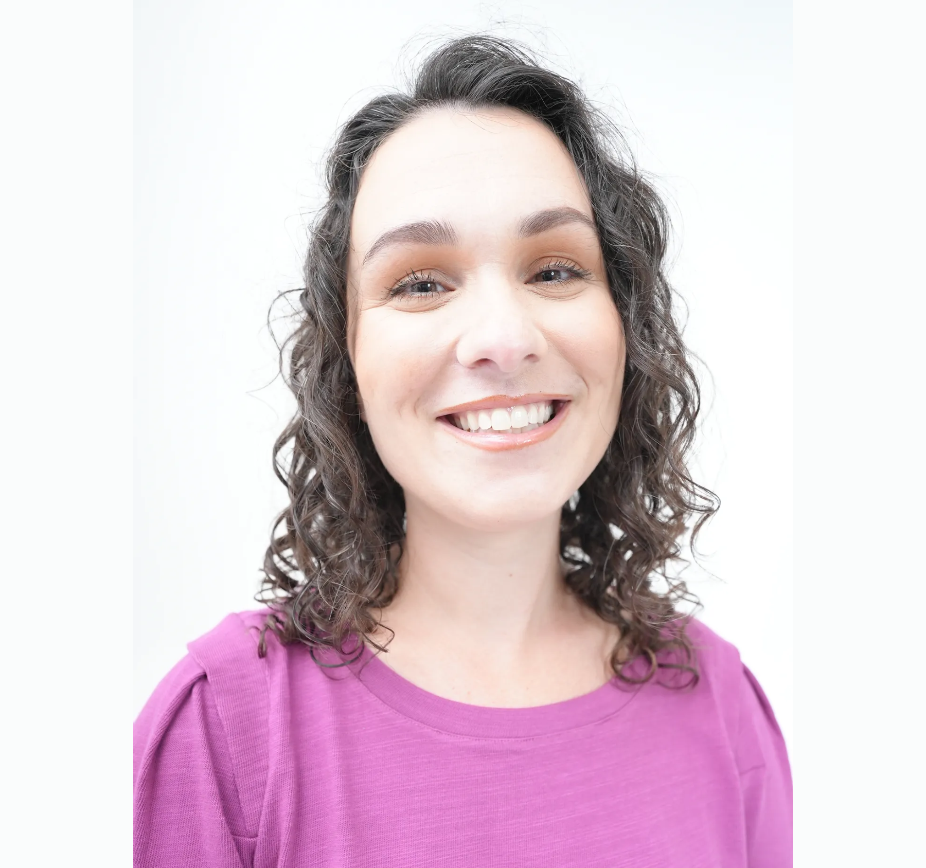 Smiling woman with curly dark hair wearing a purple top against a white background.