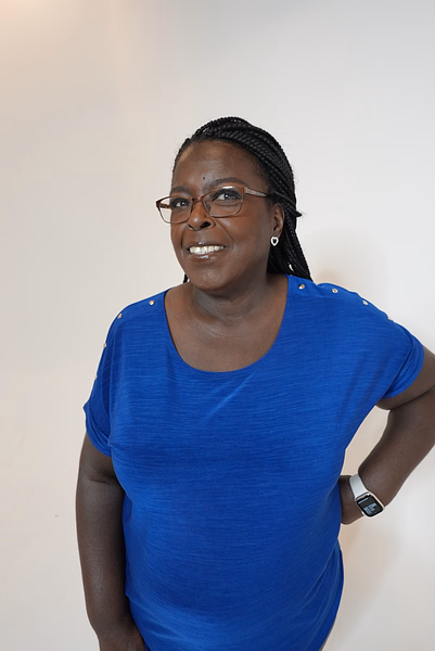 Smiling middle-aged woman wearing glasses and a blue shirt, standing against a plain white background.