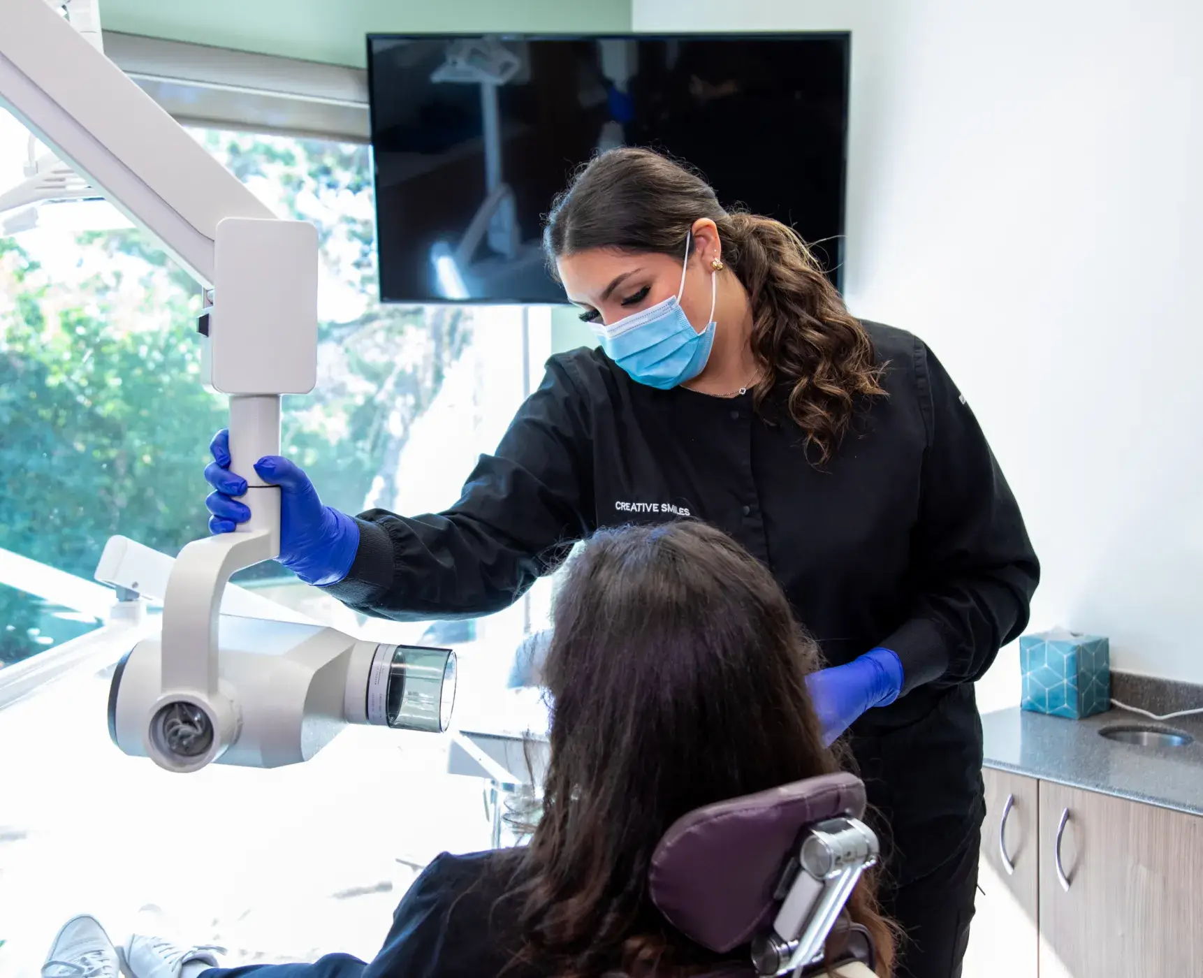 Dentist wearing a mask and gloves examines a patient's teeth using dental equipment.