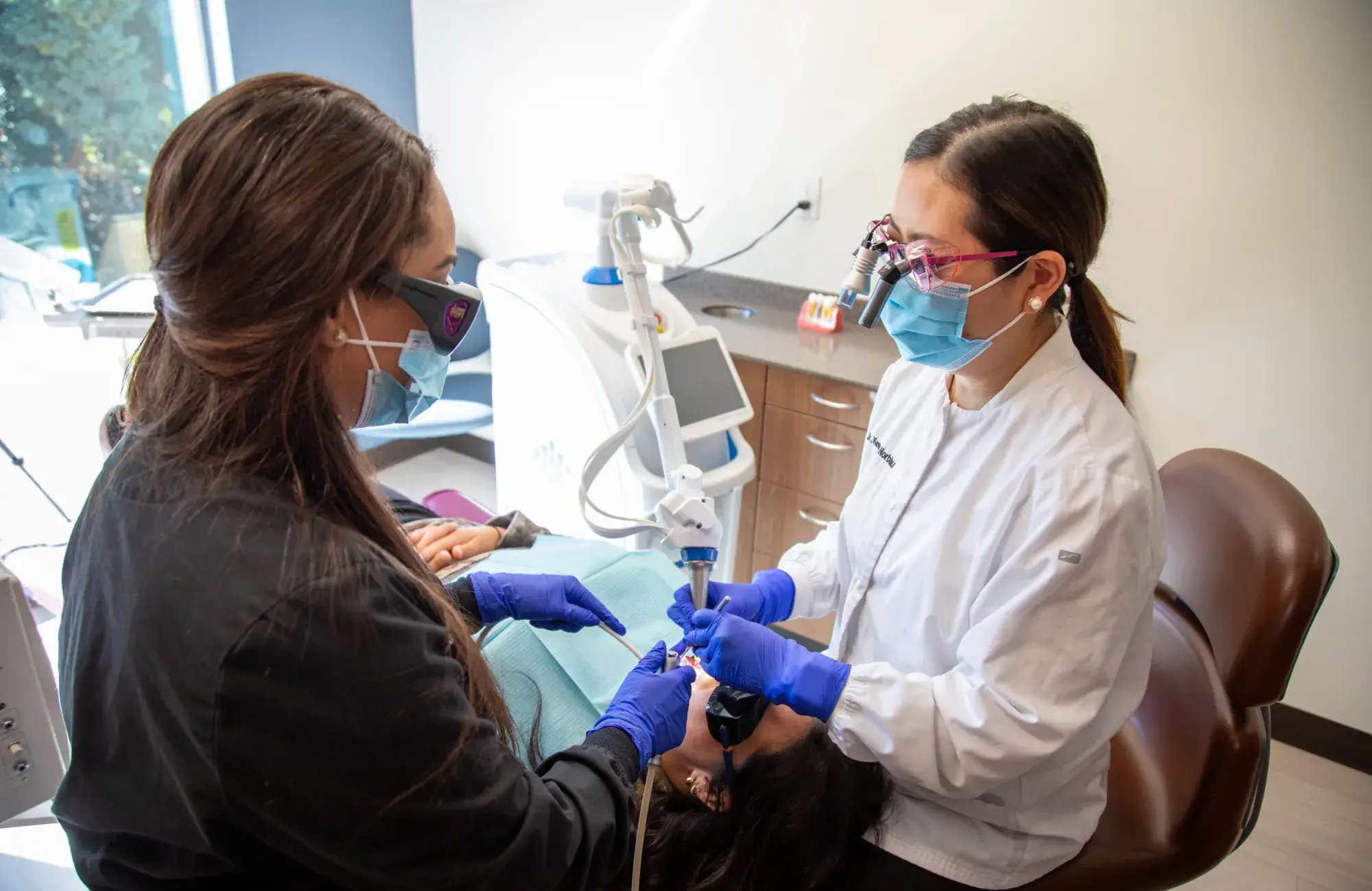 Two dental professionals in masks and gloves perform a procedure on a patient in a dental office.