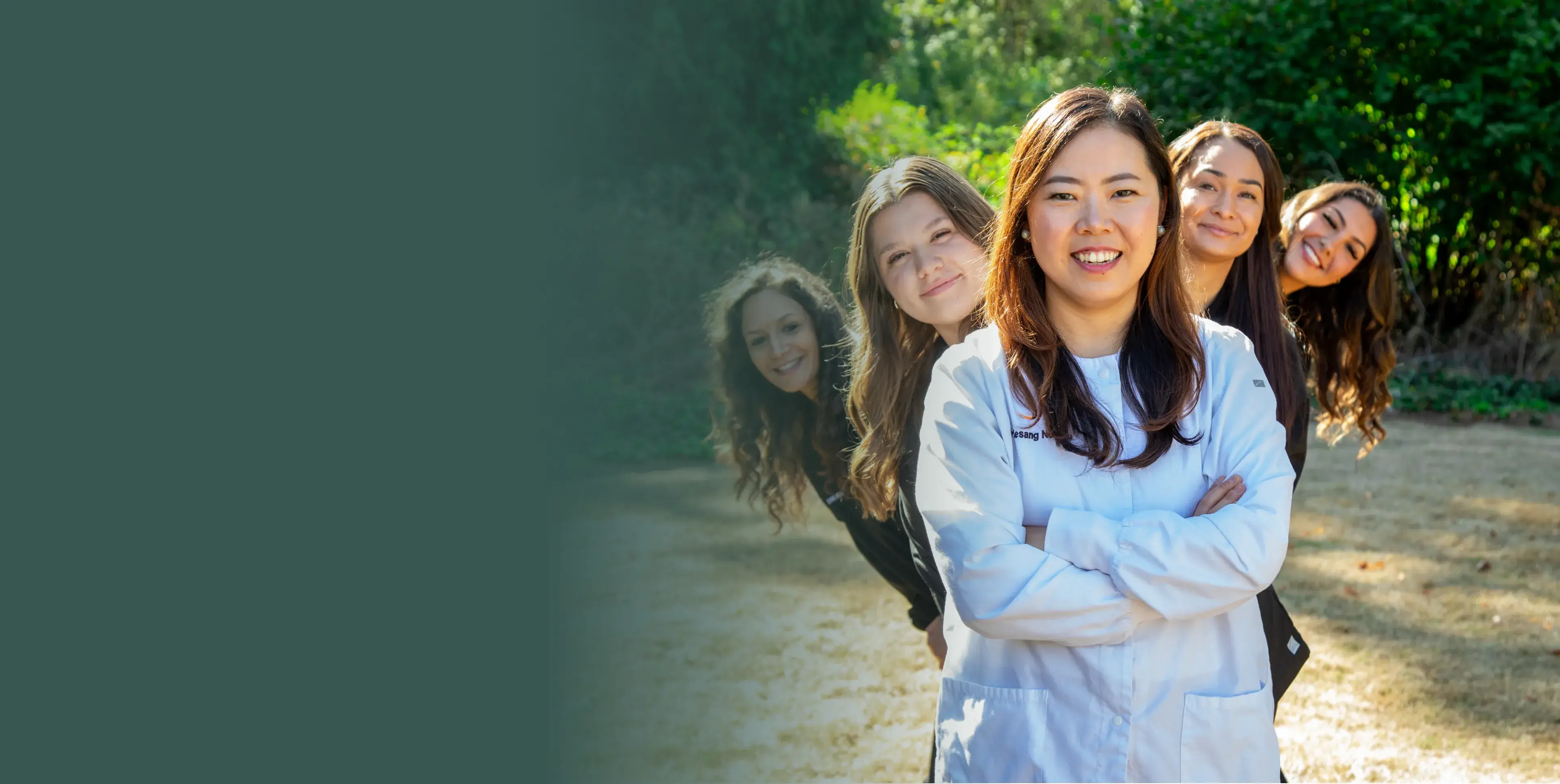 A woman in a lab coat stands smiling, with four people peeking from behind her outside.