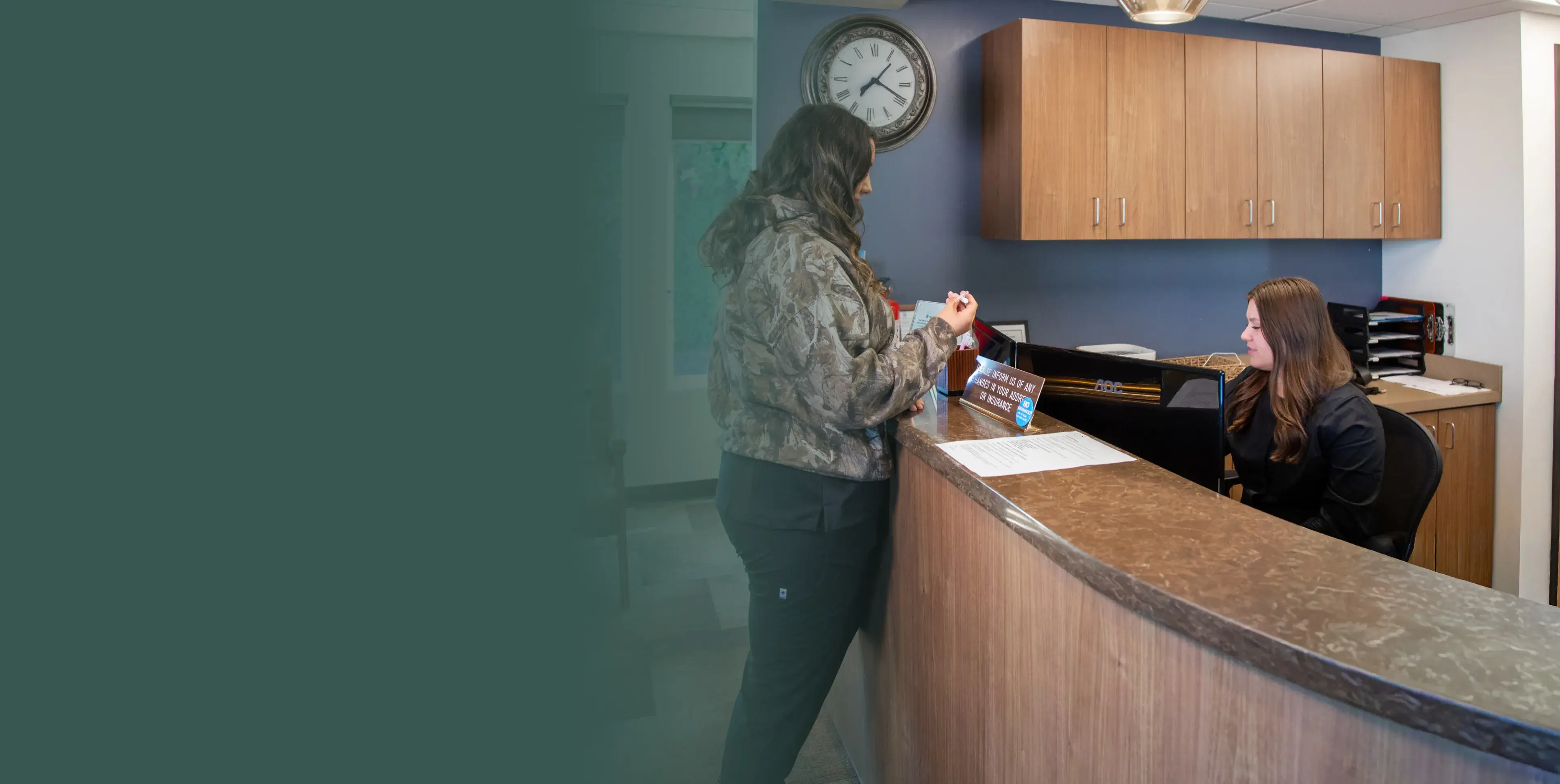 A woman stands at a reception desk, talking to a receptionist seated behind the counter.