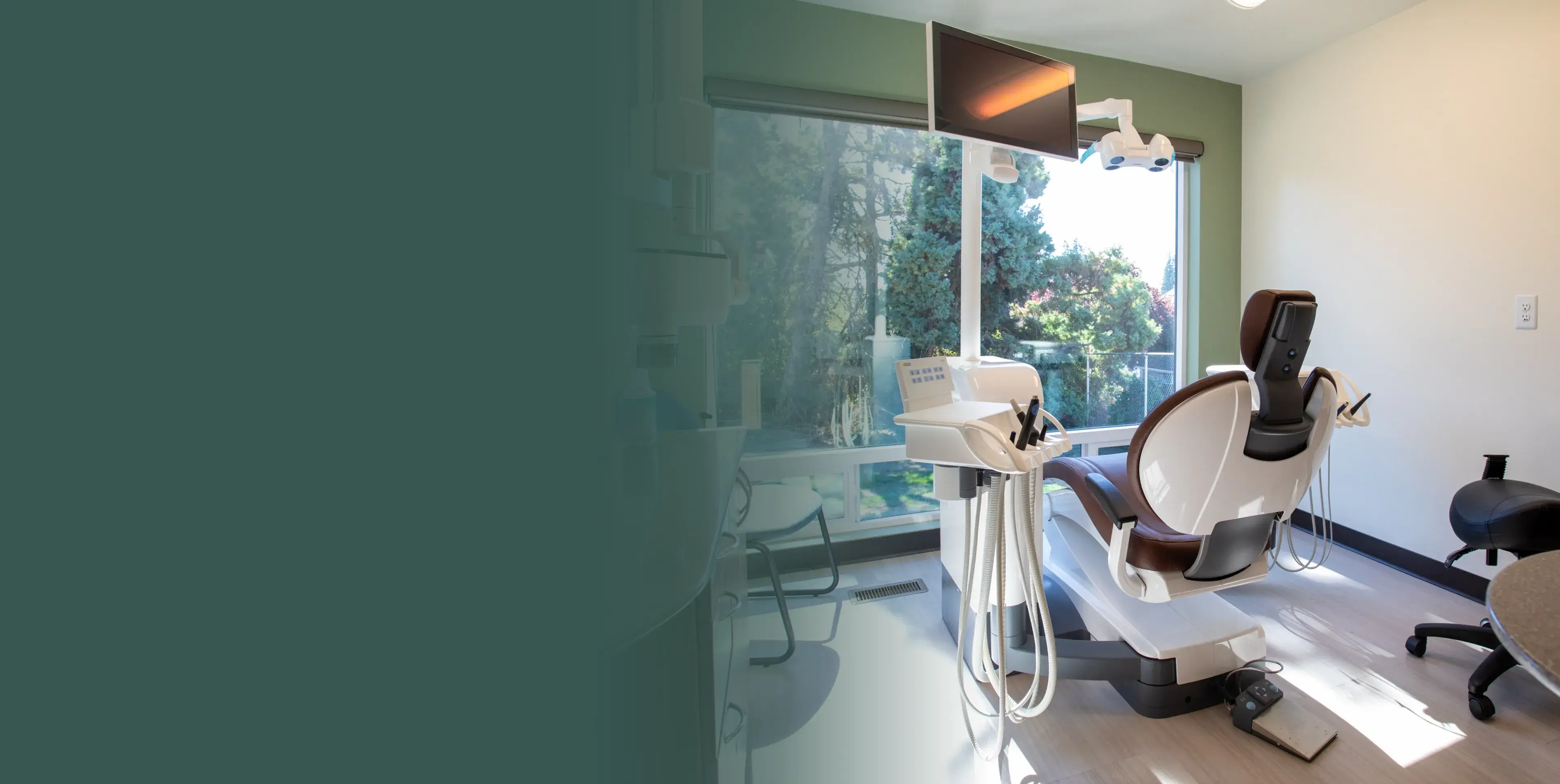 Dental examination room with a dentist chair, overhead light, and a monitor. Bright window view.