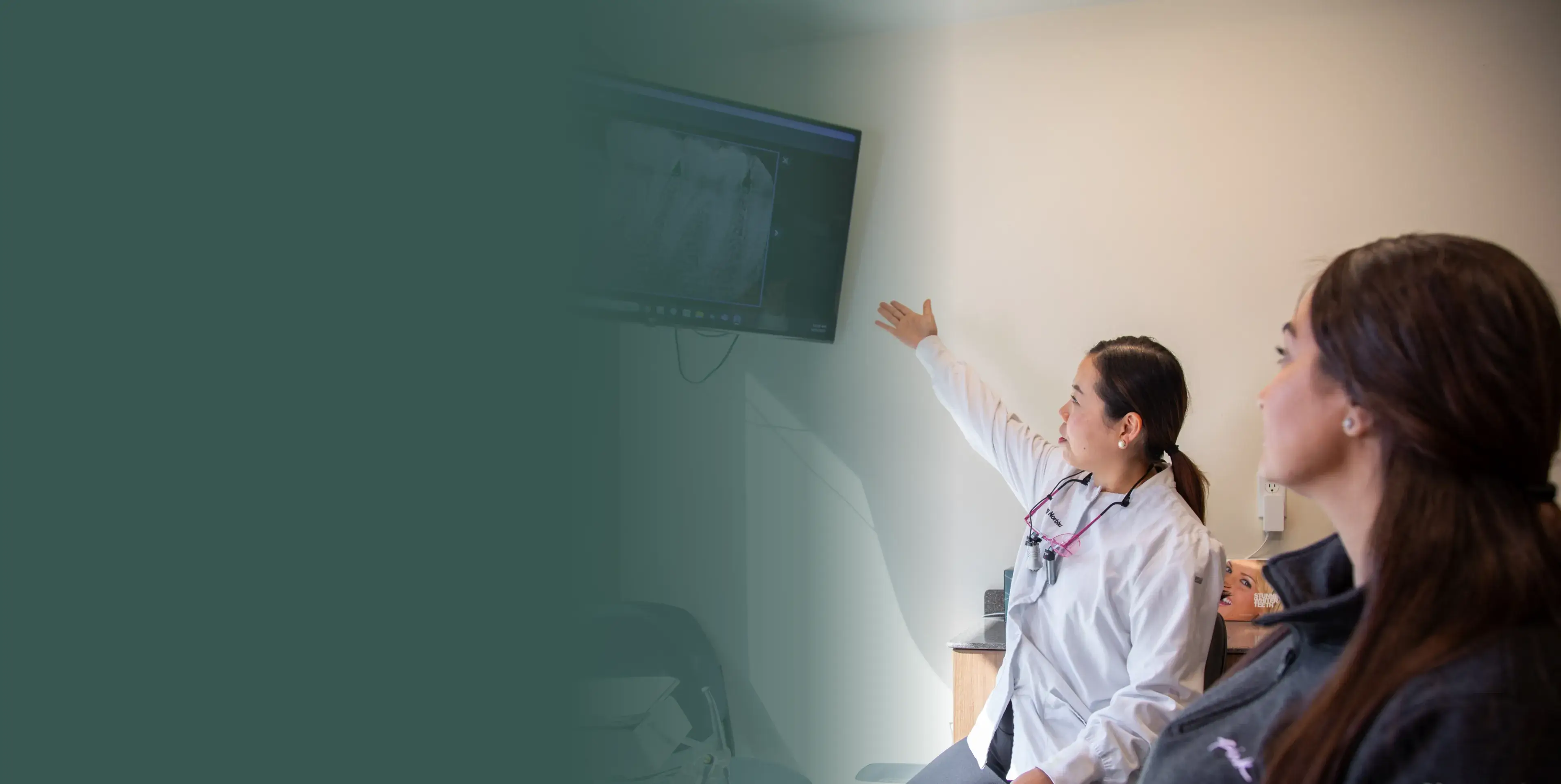 A dentist points at an x-ray on a screen while explaining to a patient.