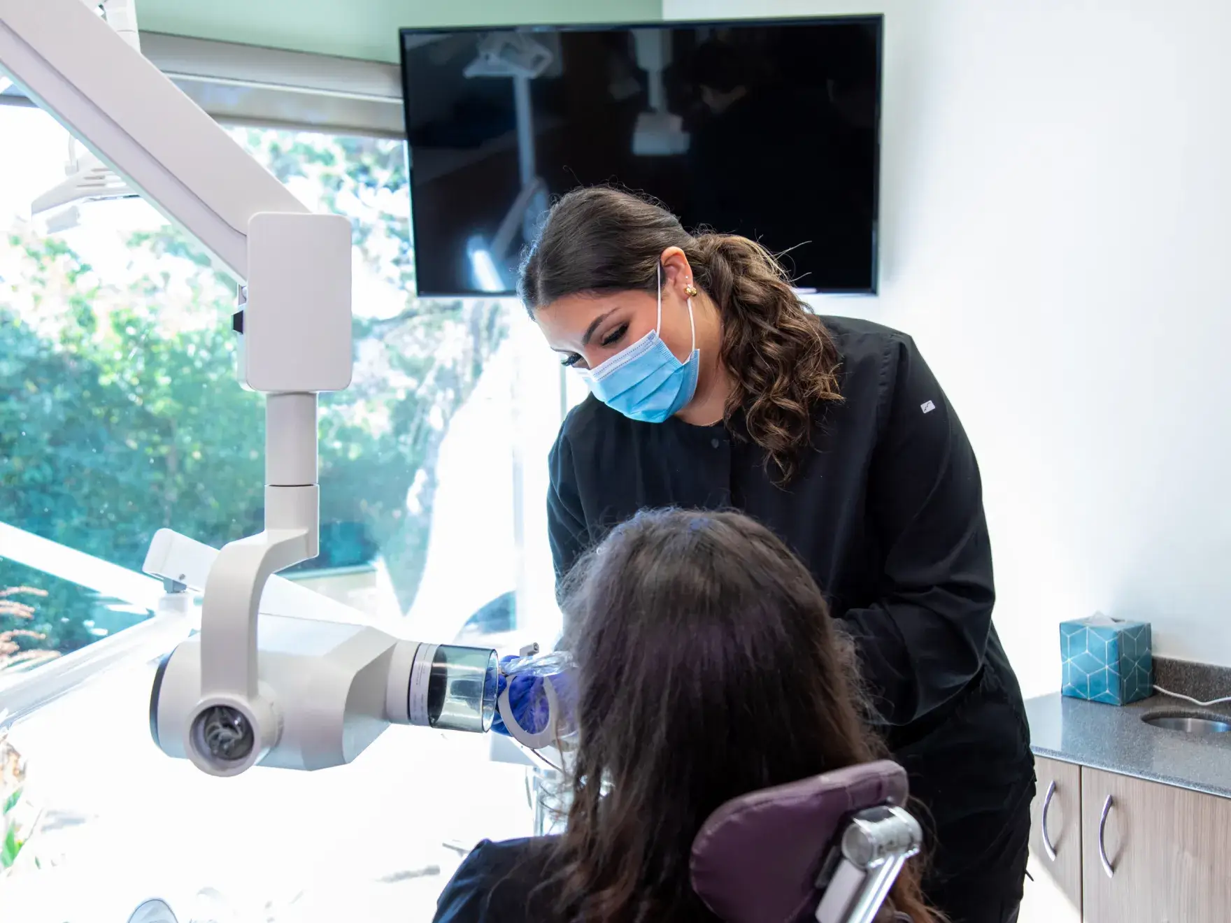 Dentist wearing a mask examines a patient's teeth using dental equipment in a clinic.
