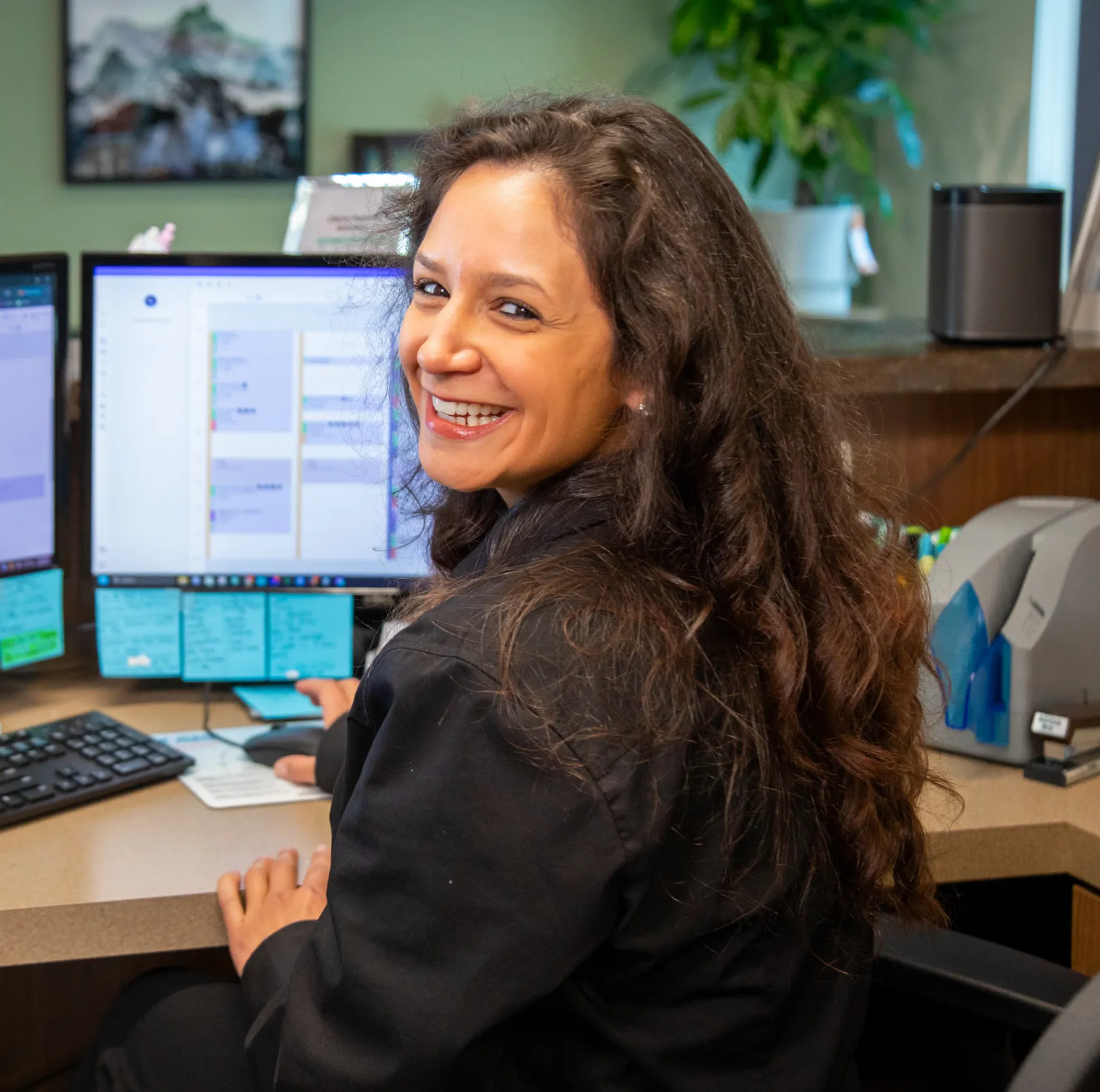 Woman sitting at a desk, smiling at the camera, with computer screens in front of her.