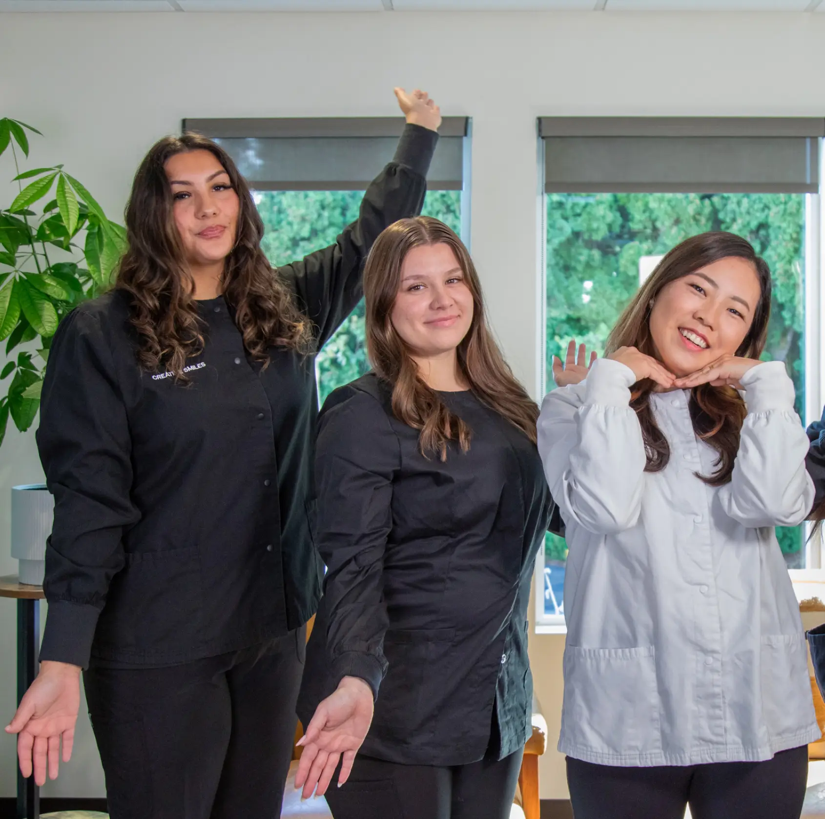 Five smiling women in casual outfits pose confidently in front of three large windows.