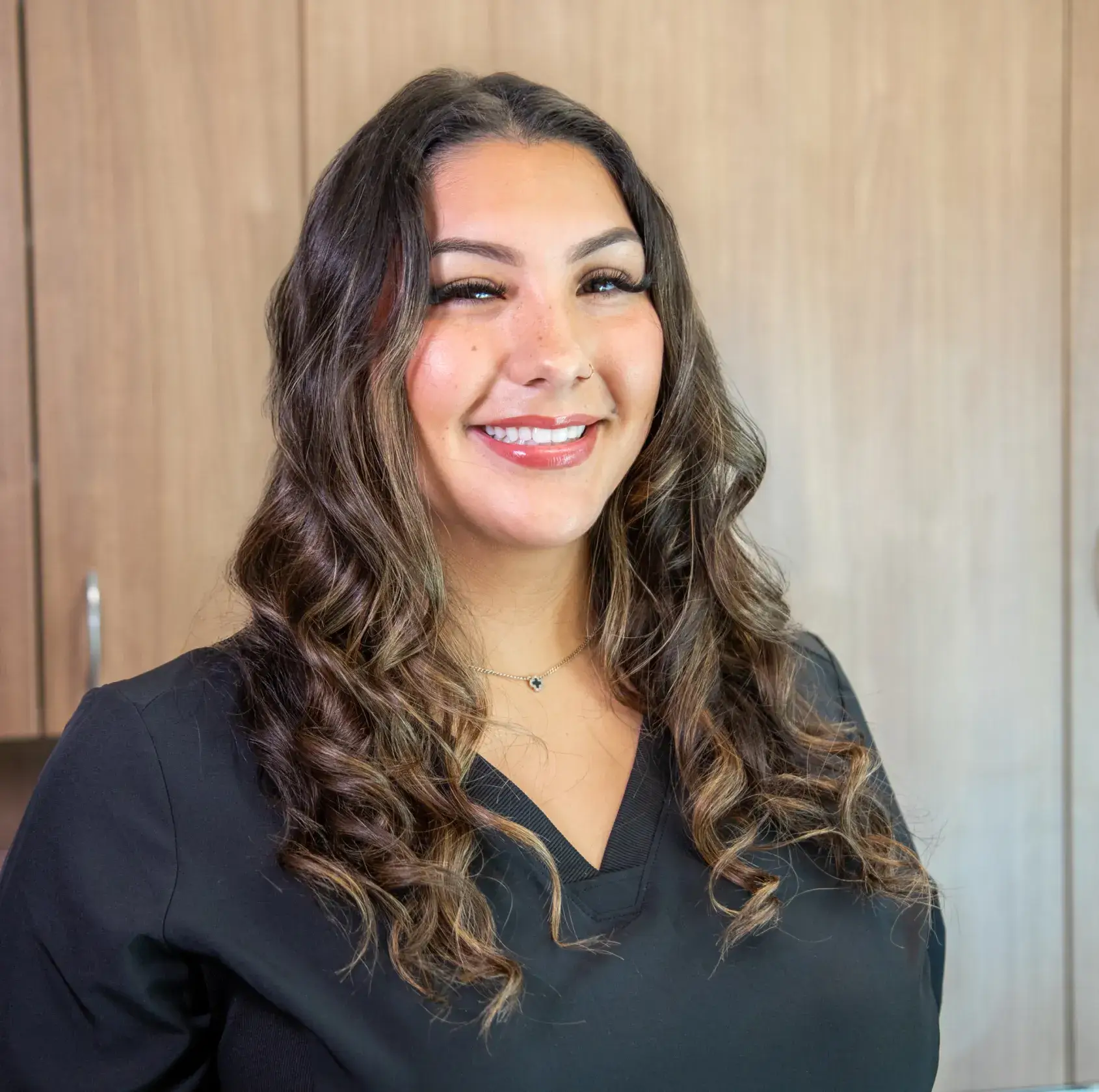 Smiling person with long, wavy hair wearing a black shirt in front of wooden cabinets.