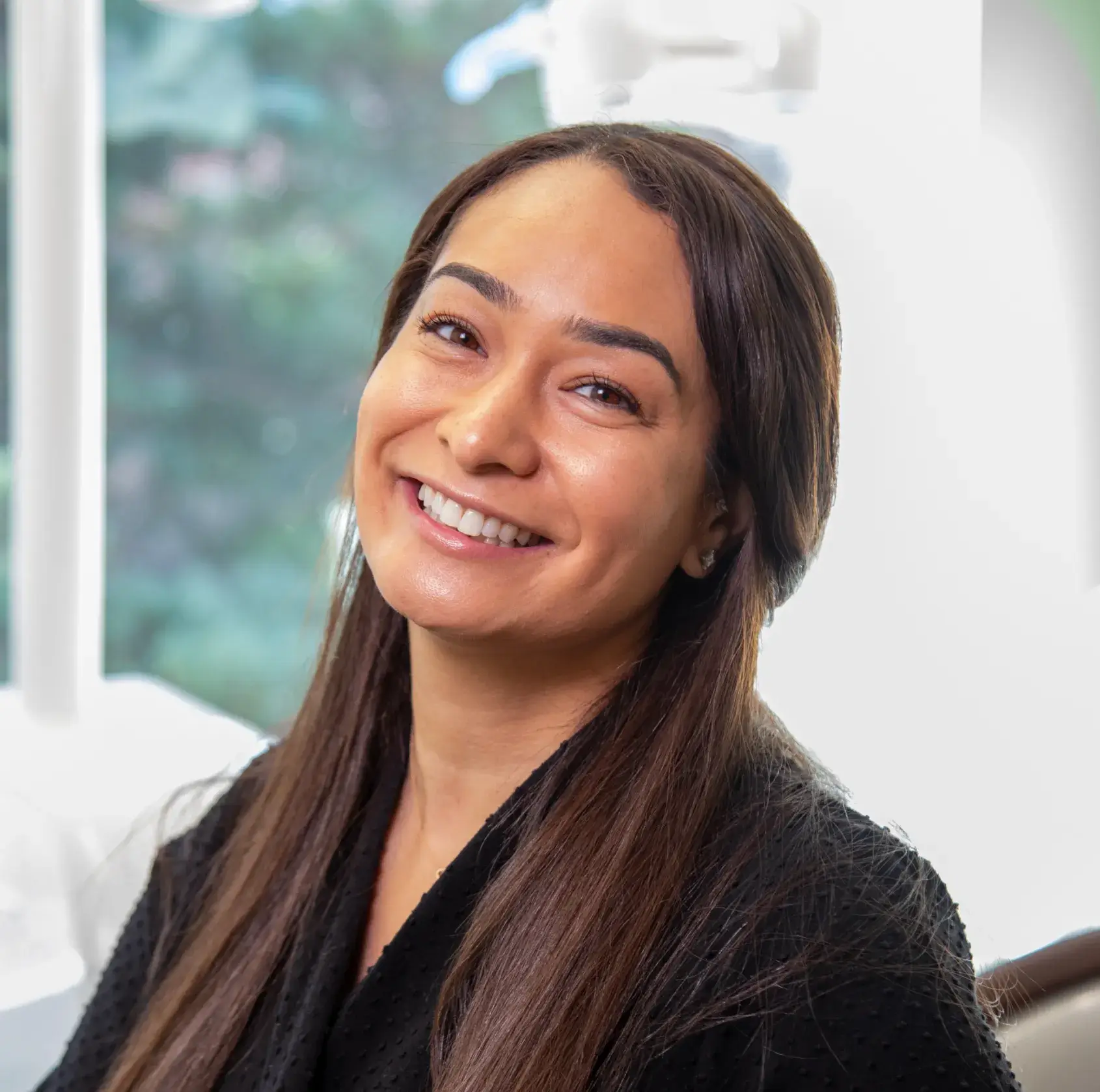 A person with long hair smiles warmly while seated indoors.