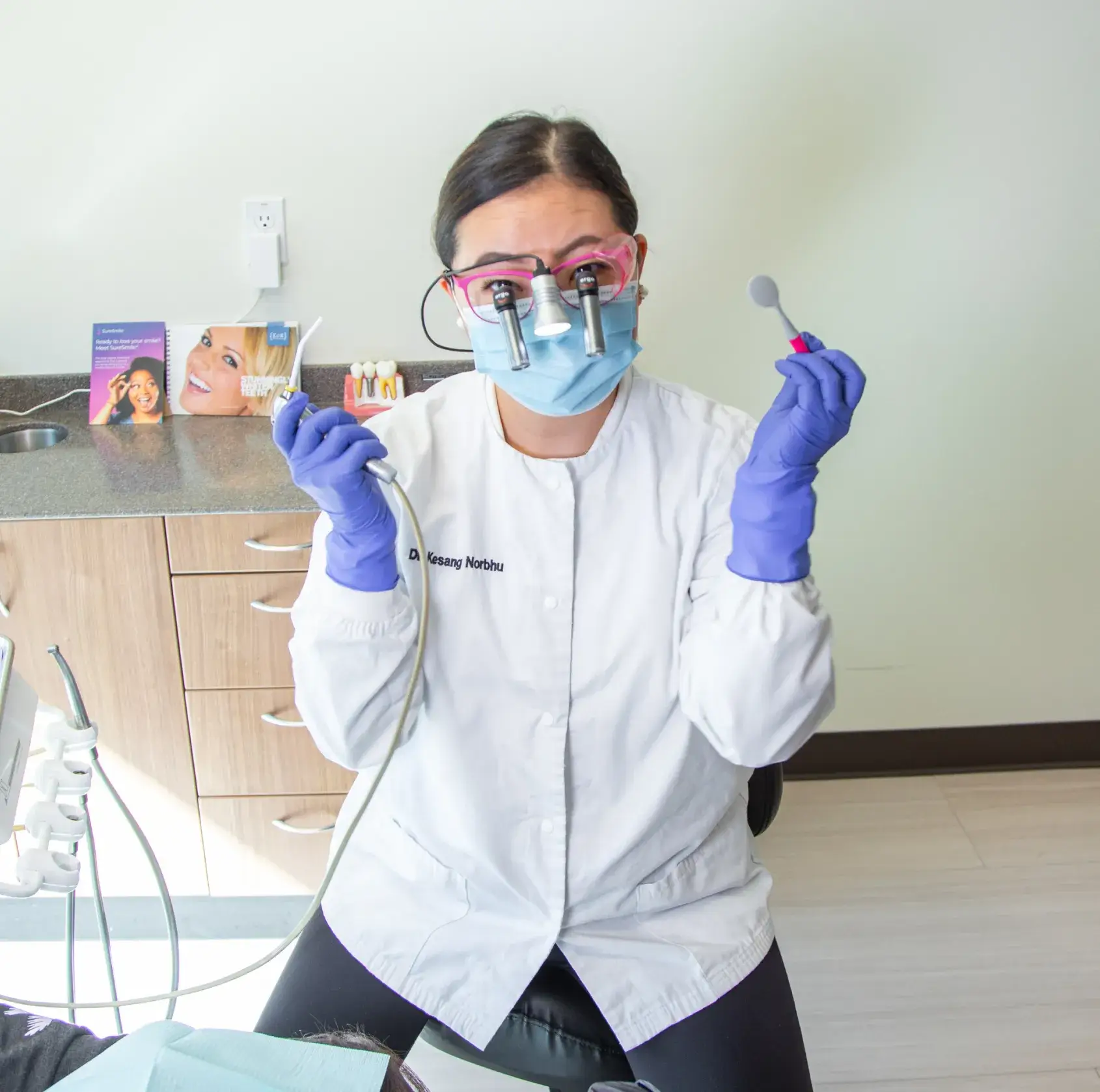 Dentist wearing a mask and gloves, holding dental tools, sitting in a clinic office.