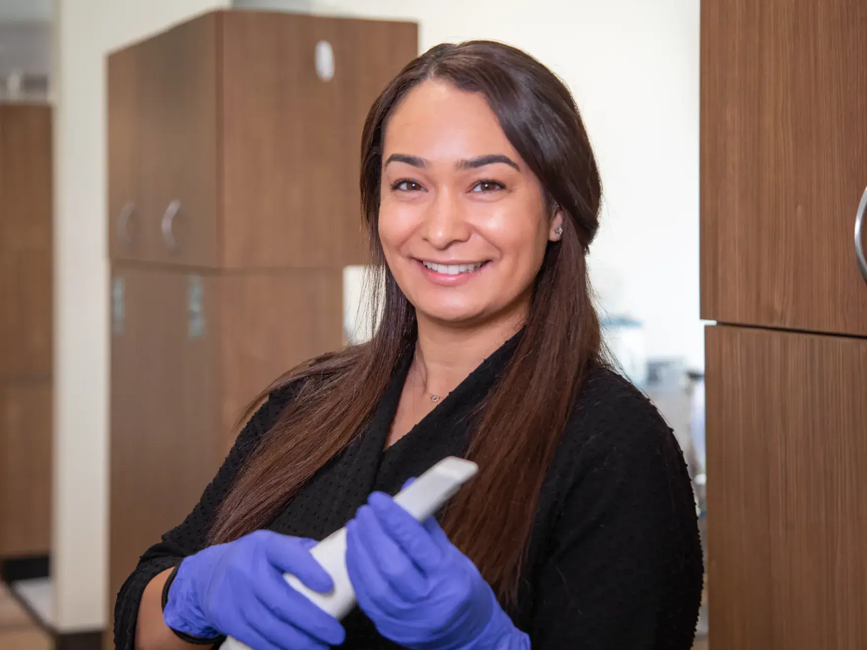 A woman in a dental office smiles while holding a dental tool, wearing blue gloves.