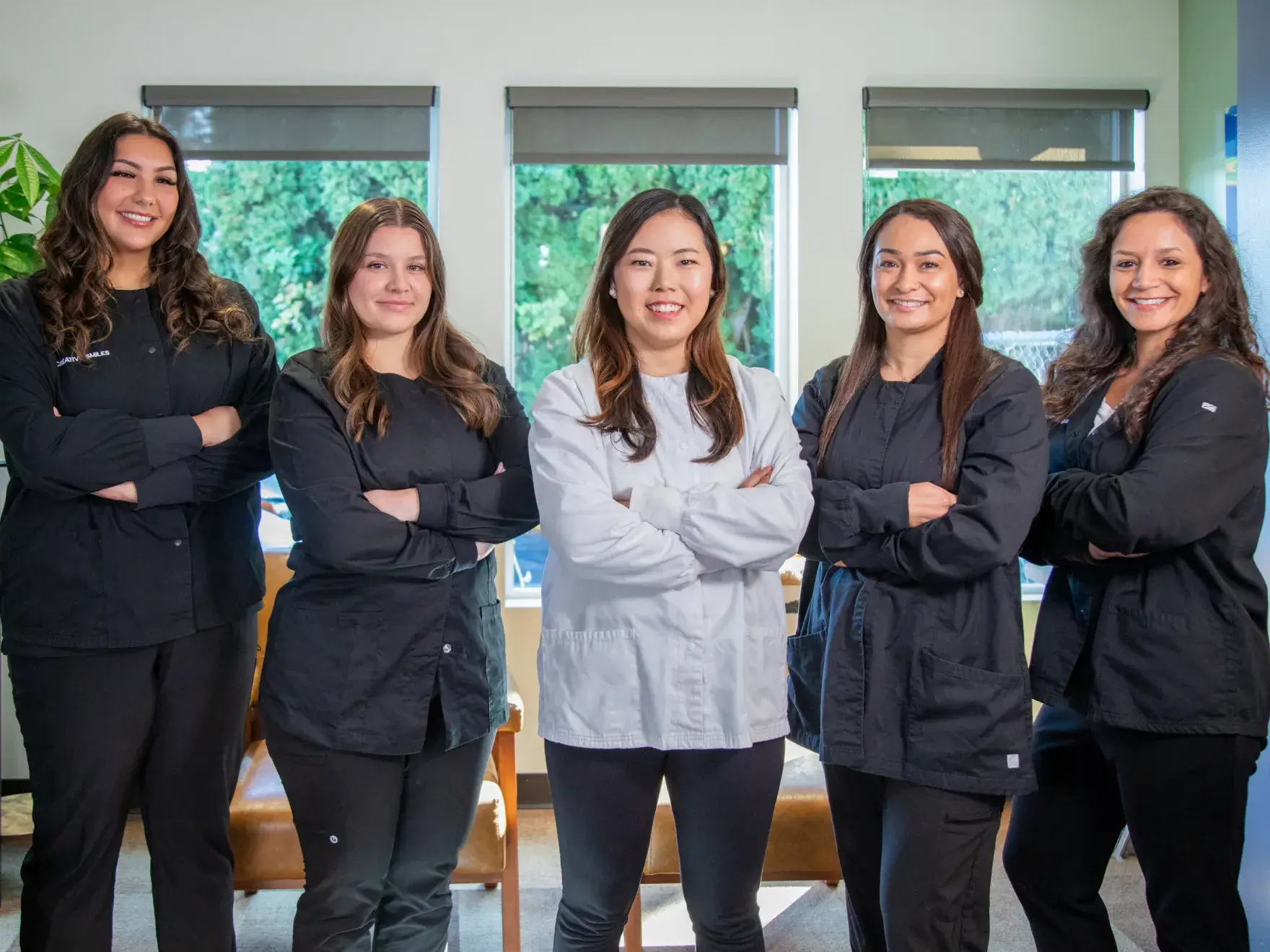 Five women standing together indoors, smiling, wearing professional attire with arms crossed.