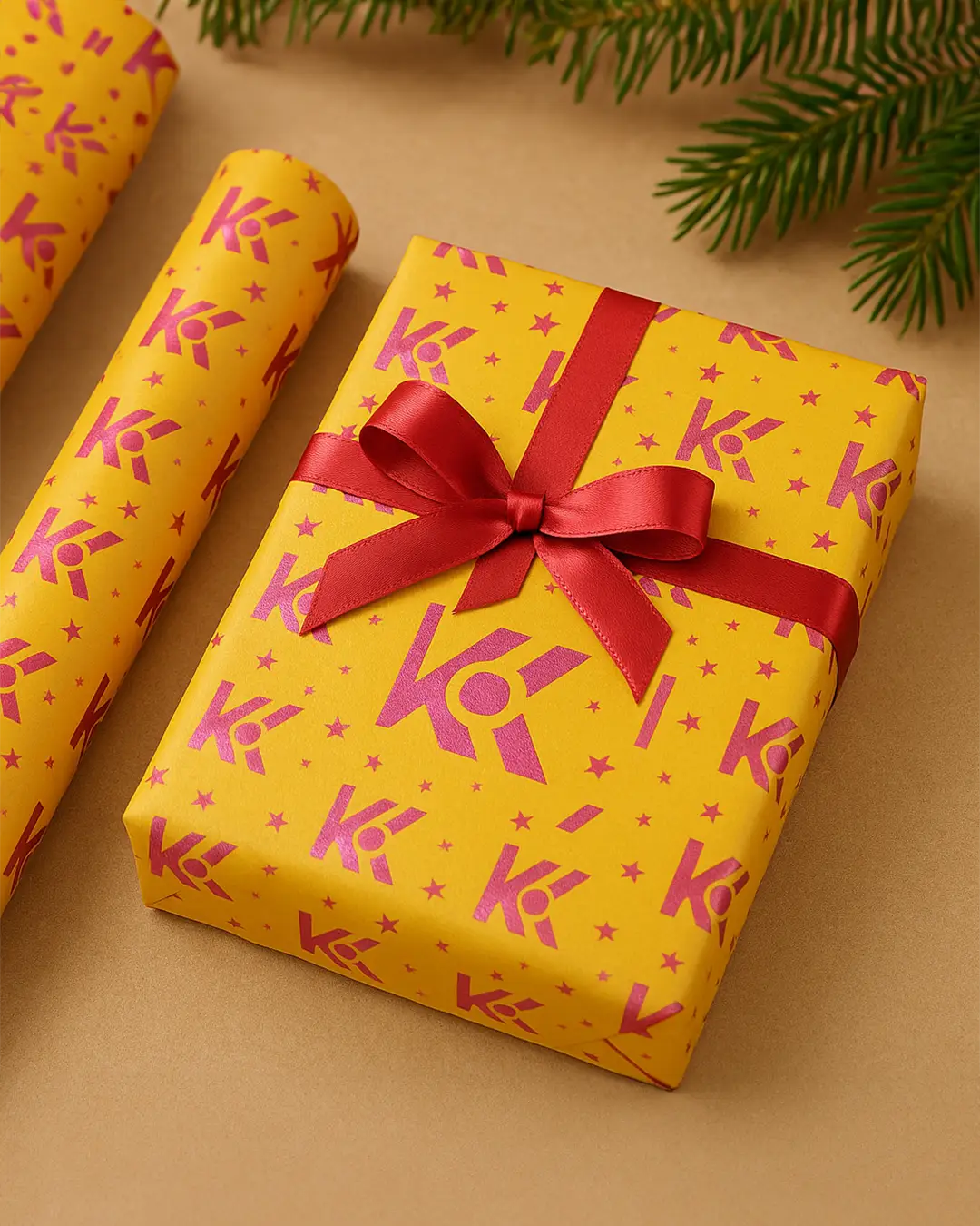 elb-rot verpacktes Geschenk mit rotem Satinband, dekoriert mit wiederholtem ‚Kimi Weber – dieKreativtuner‘-Markenlogo in Pink. Daneben liegt passendes Geschenkpapier mit gleichem Muster, auf einem warmen, beigefarbenen Hintergrund und Tannenzweig im Bildrand.