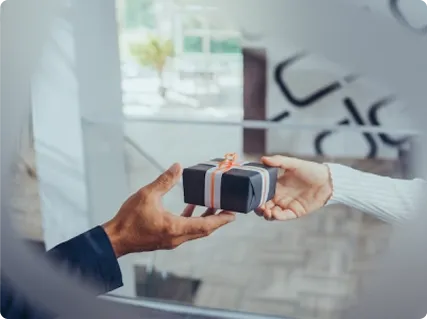 Eine Hand übergibt eine kleine schwarze Geschenkbox mit grauem Band und oranger Schleife an eine andere Hand.