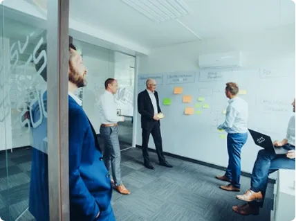 Vier Geschäftsmänner im Meetingraum, die gemeinsam an einer Wand mit Haftnotizen und Diagrammen arbeiten.