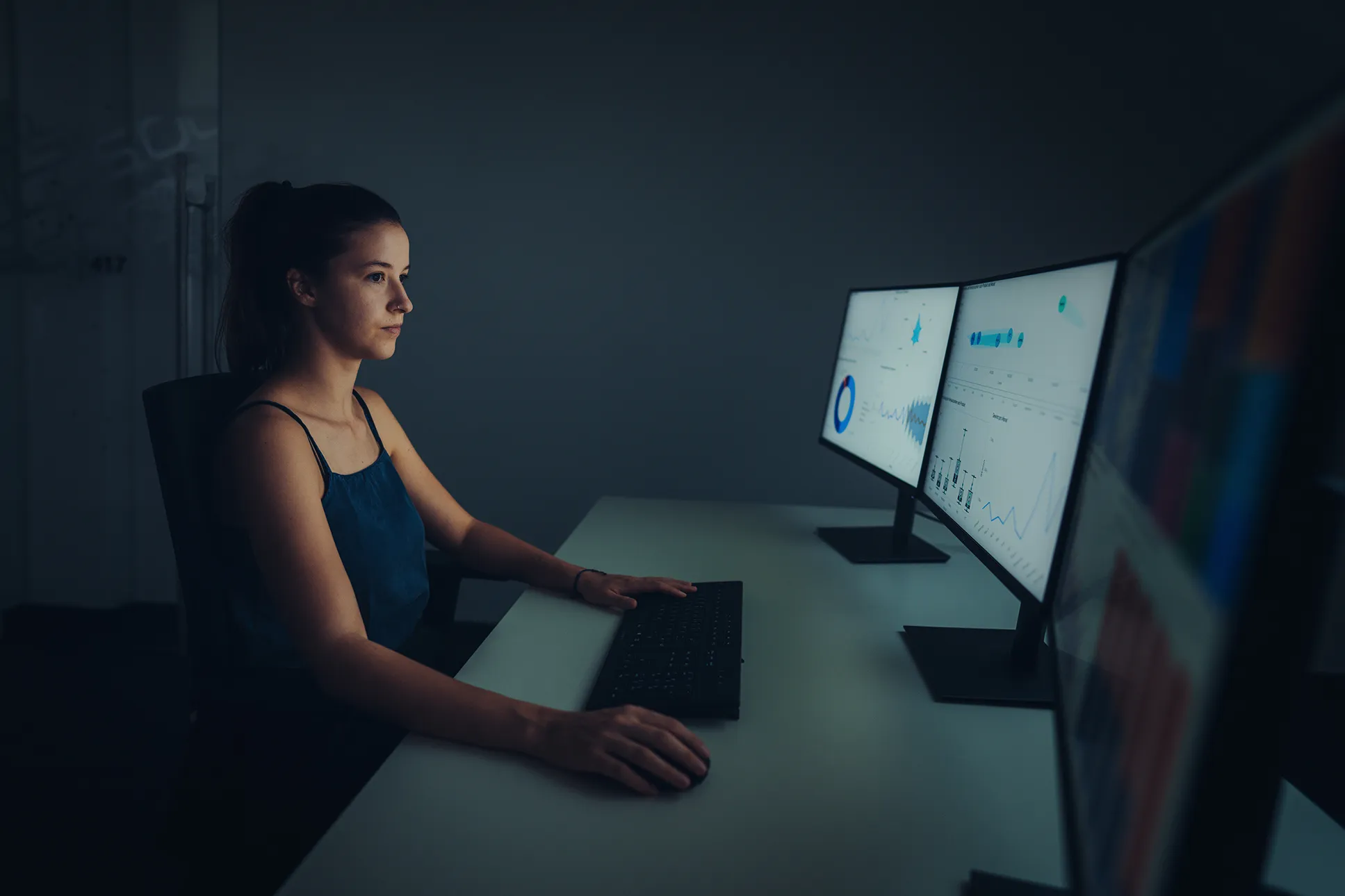 Frau arbeitet konzentriert an einem Schreibtisch mit drei Monitoren, die Daten und Diagramme anzeigen.