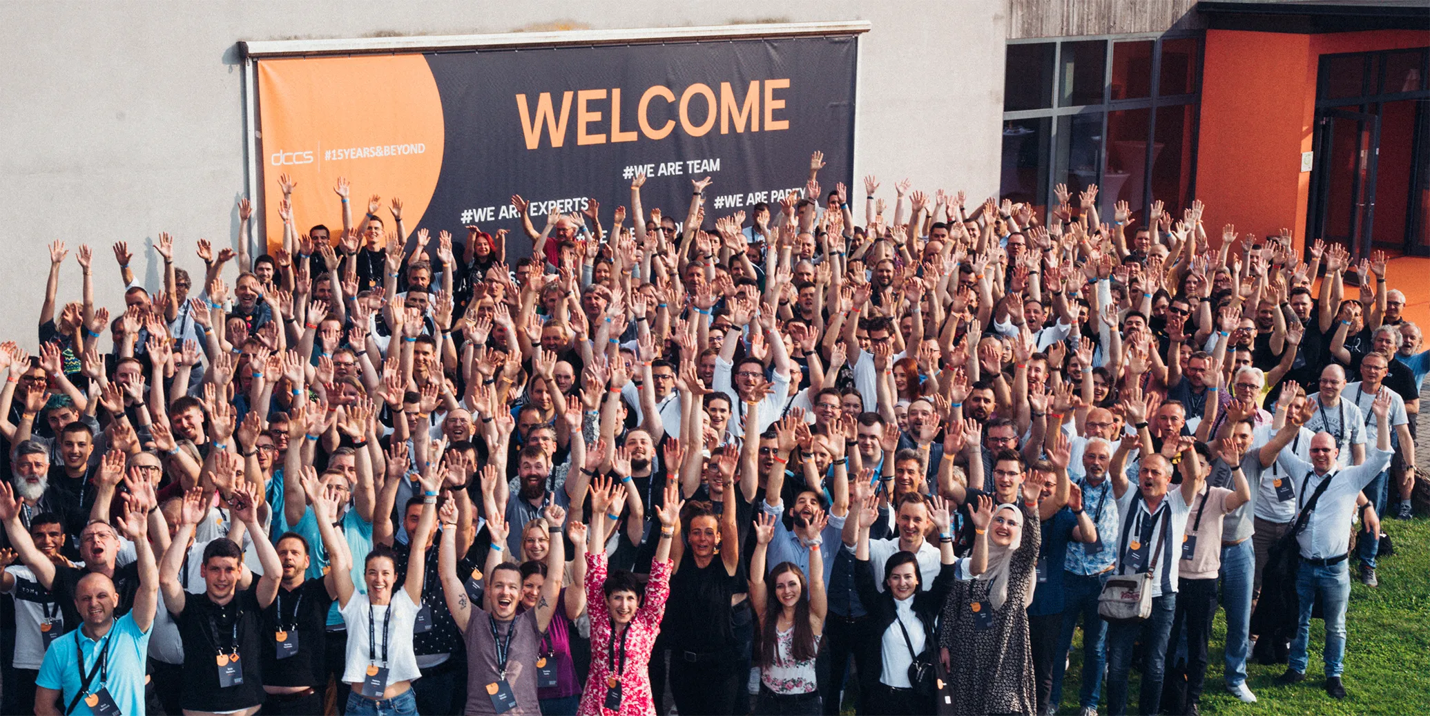 Große Gruppe von Menschen, die lächelnd die Hände vor einem Schild mit der Aufschrift ‚WELCOME‘ hochheben.