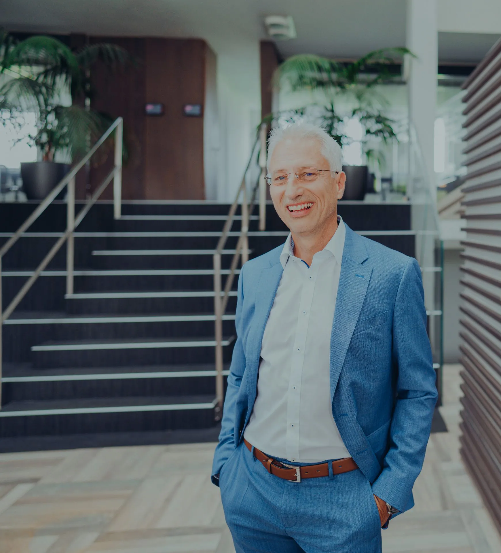 Älterer Mann in blauem Anzug mit weißem Hemd steht vor einer modernen Treppe und lächelt.