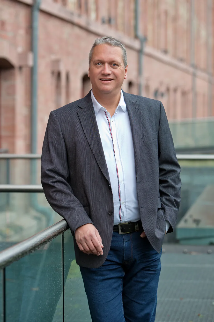 Mann in grauem Sakko und weißem Hemd lehnt lächelnd an Glasgeländer vor einem Backsteingebäude.