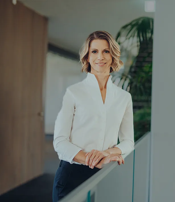 Frau mit kurzen blonden Haaren in weißer Bluse lehnt an einem Geländer in einem modernen Büro.