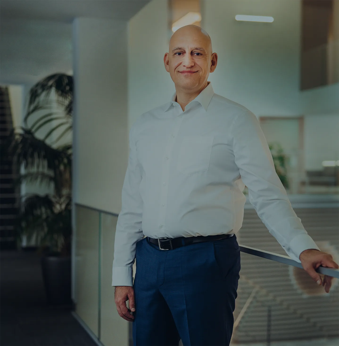 Lächelnder Mann mit weißem Hemd und blauer Hose lehnt an einem Geländer in einem modernen Bürogebäude.