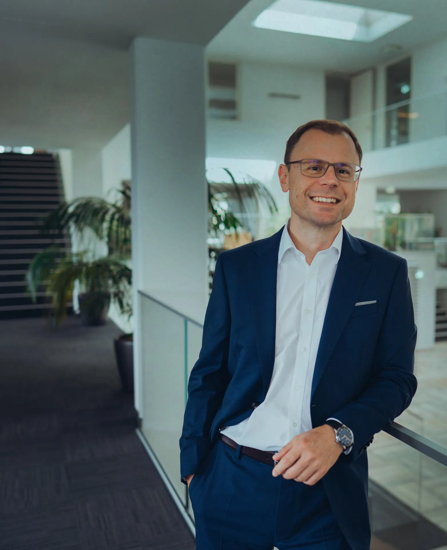 Ein lächelnder Mann in einem blauen Anzug und weißem Hemd lehnt sich in einem modernen Bürogebäude an ein Glasgeländer.