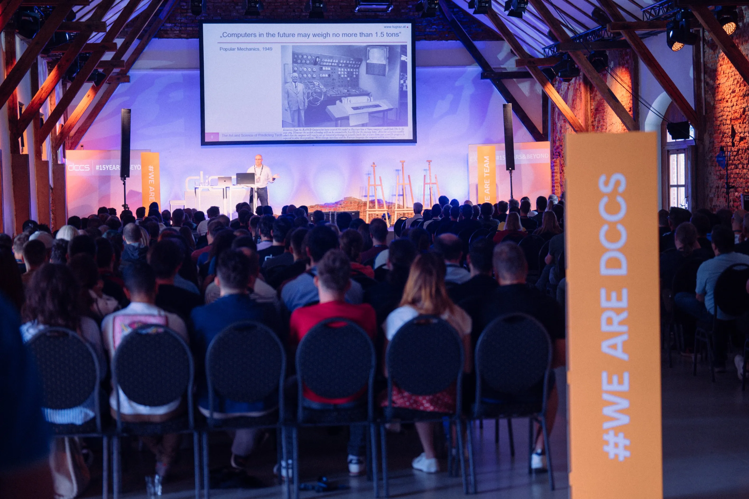 Vortragender präsentiert vor einem großen Publikum in einem Saal mit sichtbaren Balken und Steinwänden, auf einer Leinwand ist ein historisches Zitat zu Computern von 1949 zu sehen.