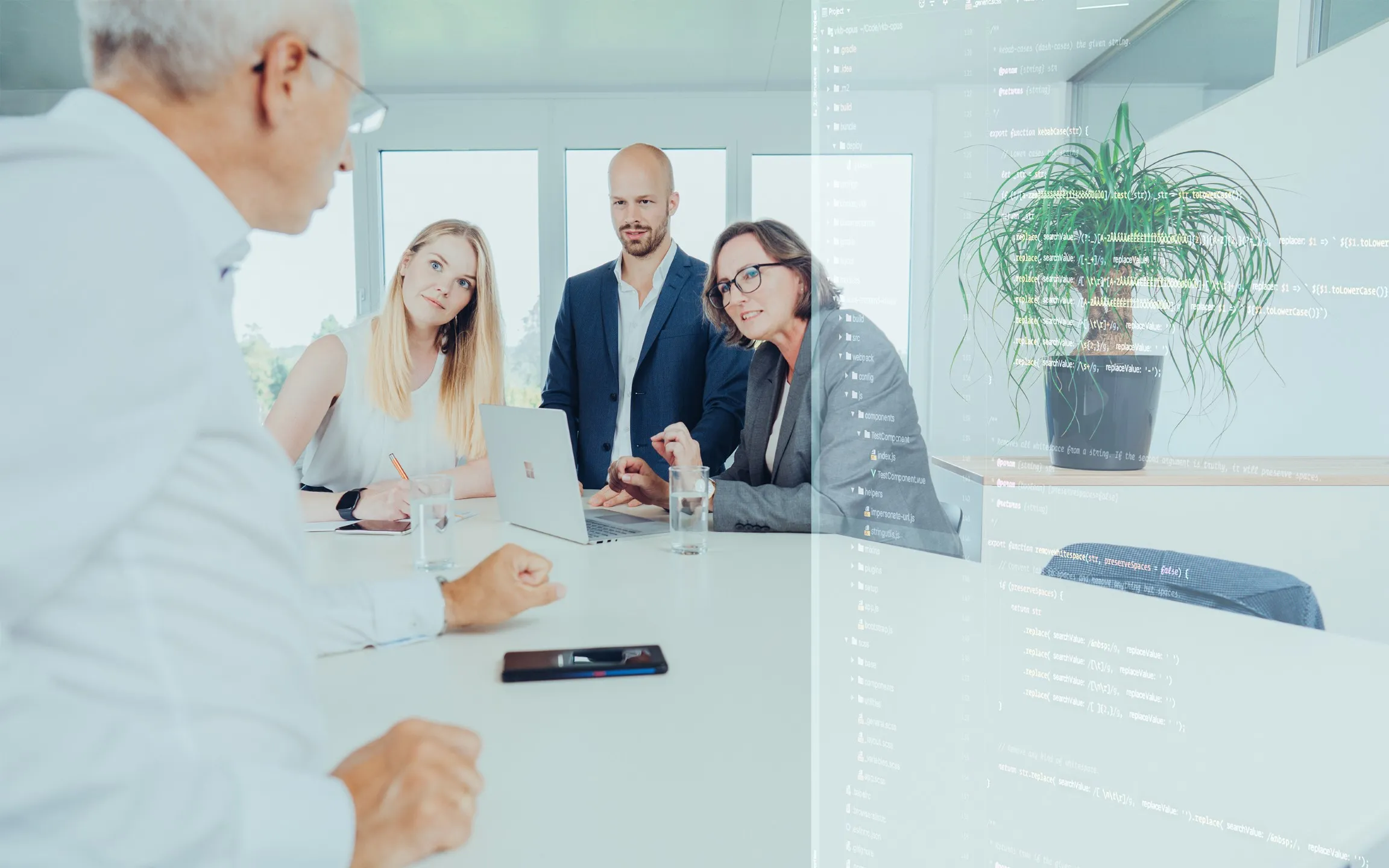 Vier Personen in einem Besprechungsraum diskutieren über einen Laptop vor einem Fenster, mit Programmiercode und einer Pflanze im Hintergrund auf einer Glaswand.