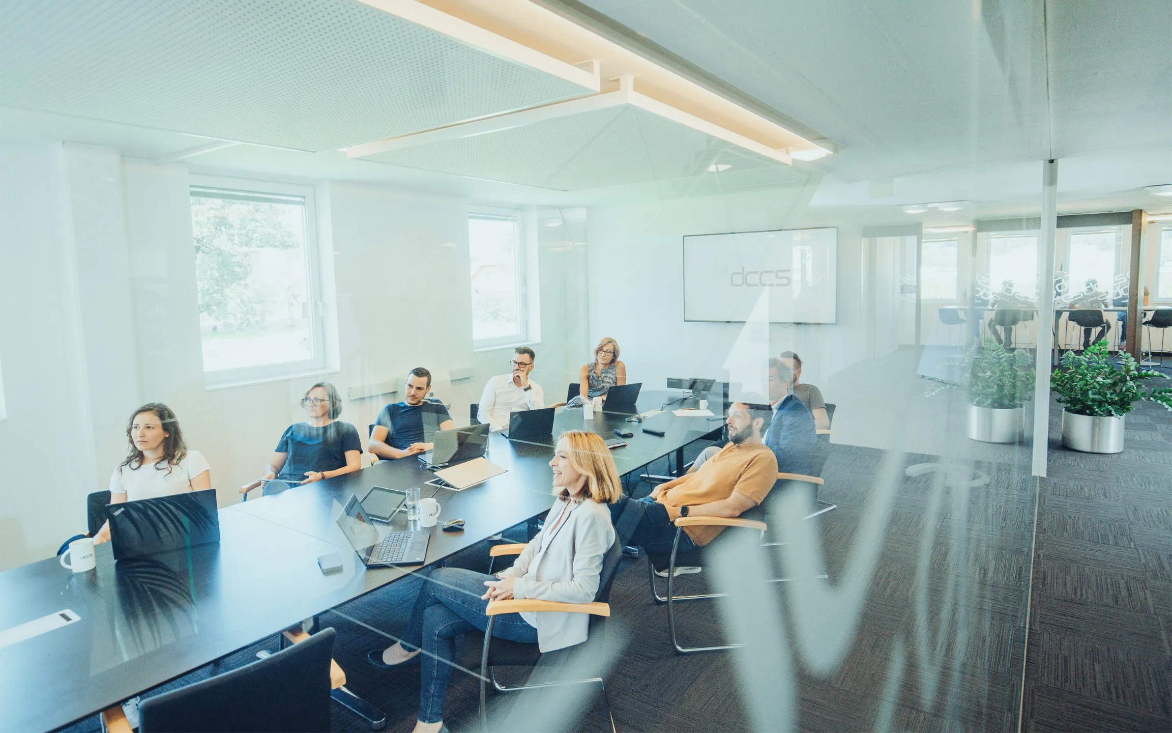 Menschen sitzen an einem Konferenztisch in einem modernen Büro, einige mit Laptops, während sie an einer Besprechung teilnehmen.