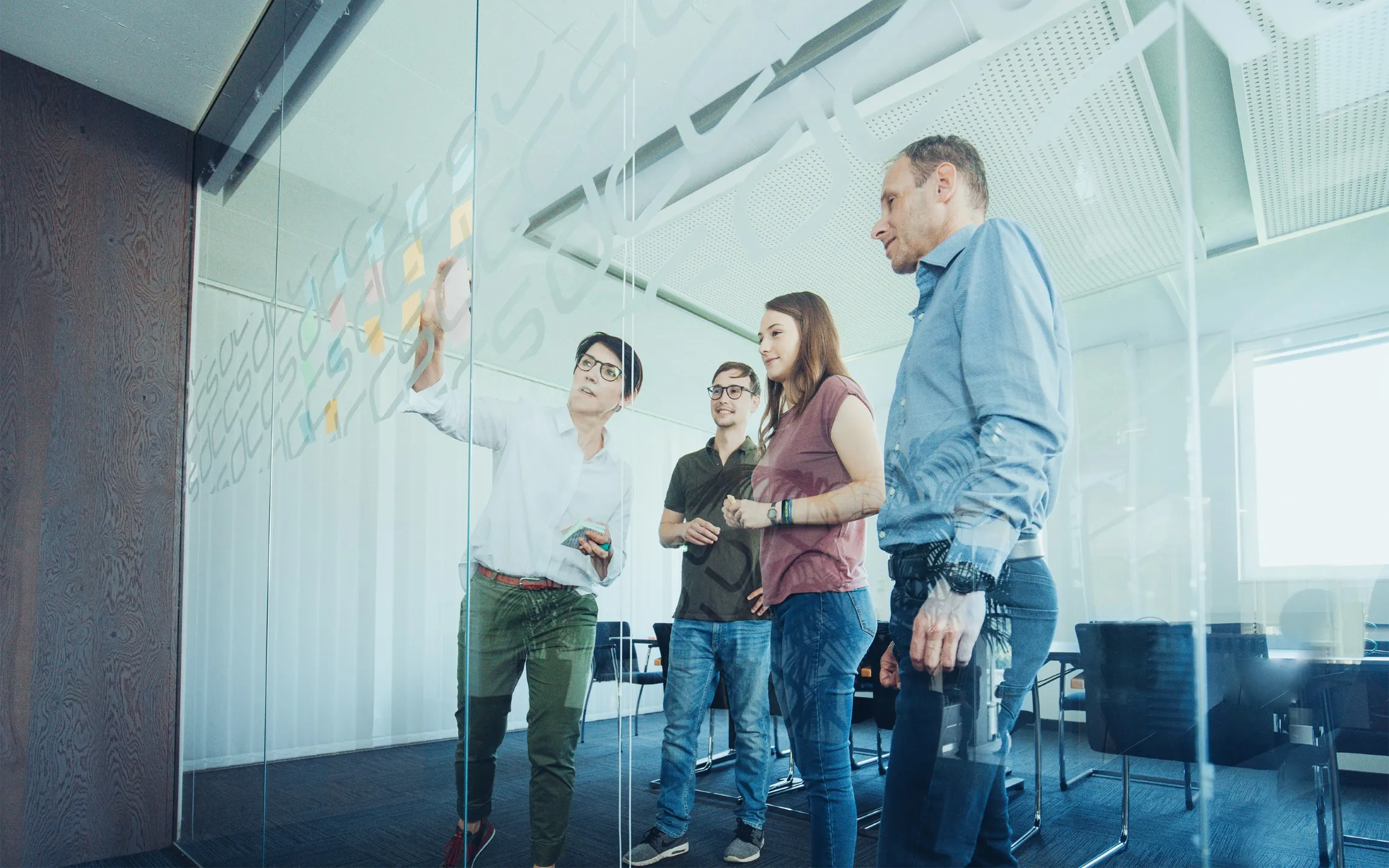 Vier Personen bei einer Team-Besprechung vor einer Glaswand mit Haftnotizen in einem modernen Büro.