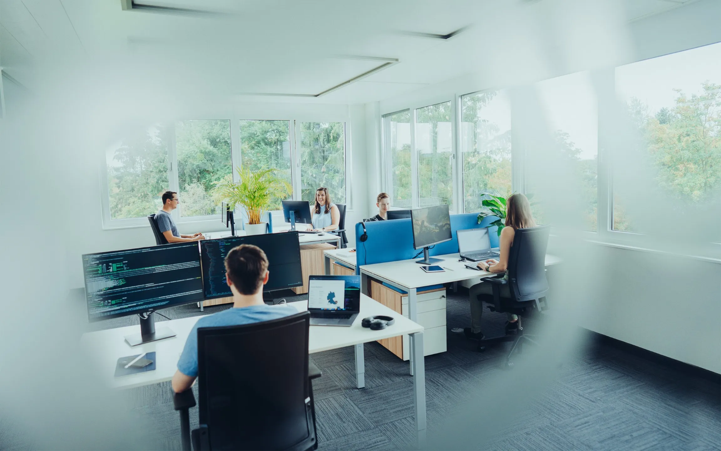 Ein modernes Büro mit fünf Personen, die an Computern an weißen Schreibtischen mit großen Fenstern und Pflanzen arbeiten.