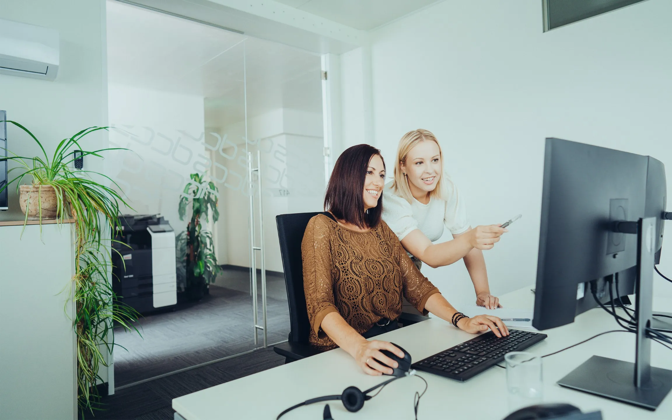 Zwei Frauen im Büro schauen gemeinsam auf einen Computermonitor, eine bedient die Maus, die andere zeigt mit einem Stift auf den Bildschirm.