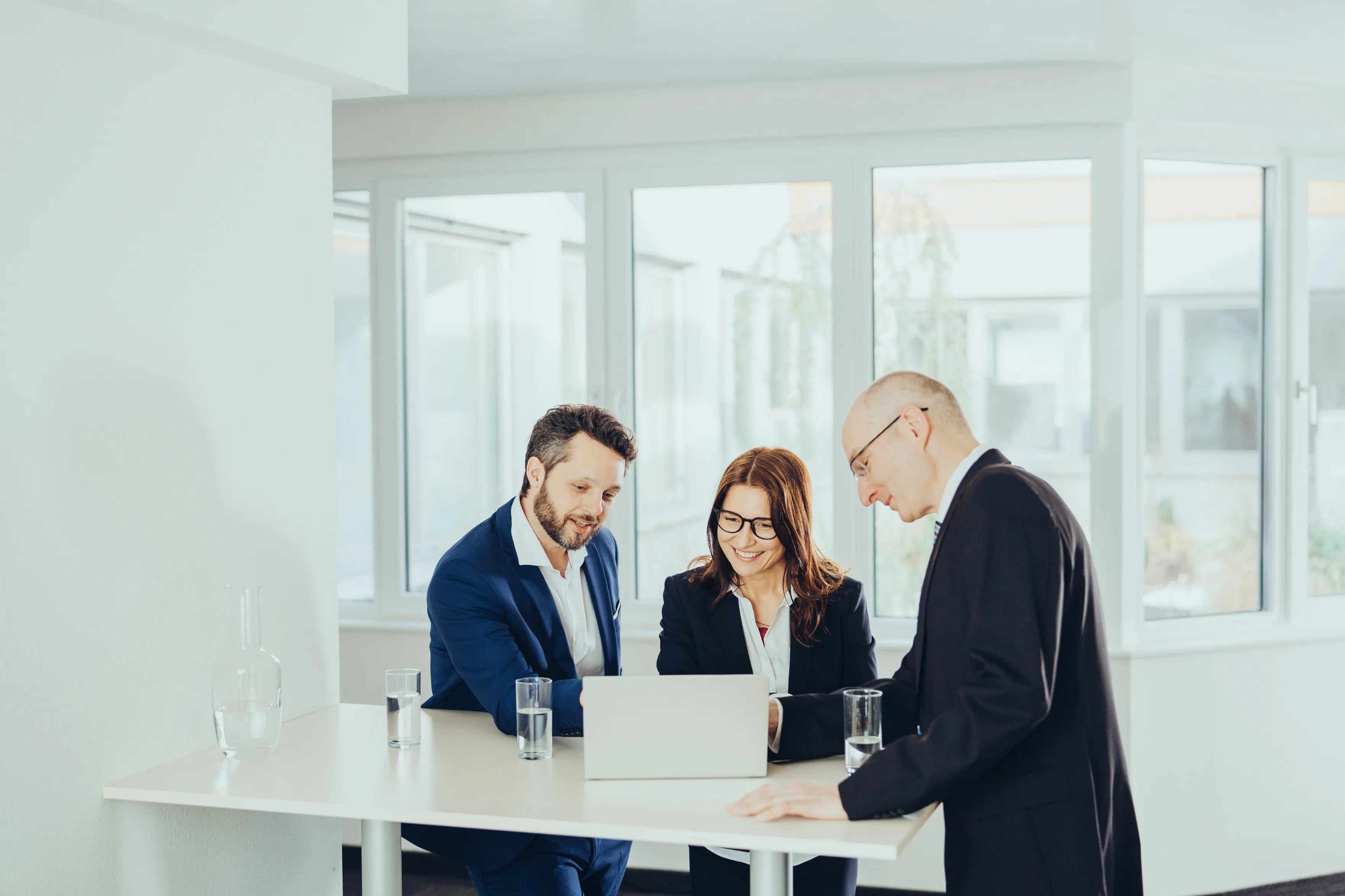 Drei beruflich gekleidete Personen an einem Stehtisch schauen gemeinsam auf einen Laptop.