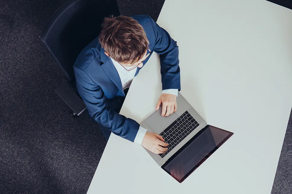 Von oben betrachteter Mann im Anzug sitzt an einem weißen Tisch und tippt auf einem Laptop.