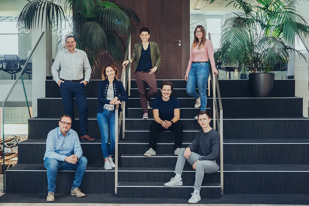 Sieben Personen in einem modernen Büro sitzen und stehen lächelnd auf einer breiten Treppe mit großen Pflanzen im Hintergrund.